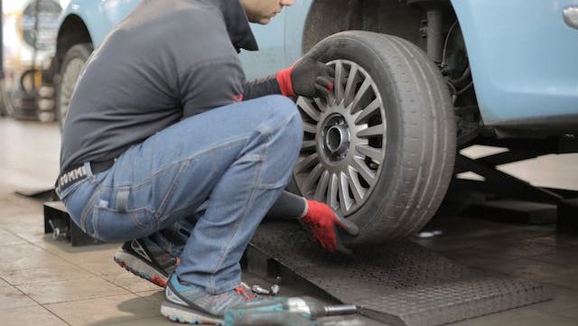 A mechanic in blue jeans and a gray shirt uses a power tool to install a tire on a light blue car in a garage. He wears red gloves, focusing intently.