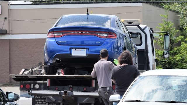 A blue Dodge car is being loaded onto a tow truck in a parking lot. Two people stand behind the truck observing. The scene conveys urgency and concern.