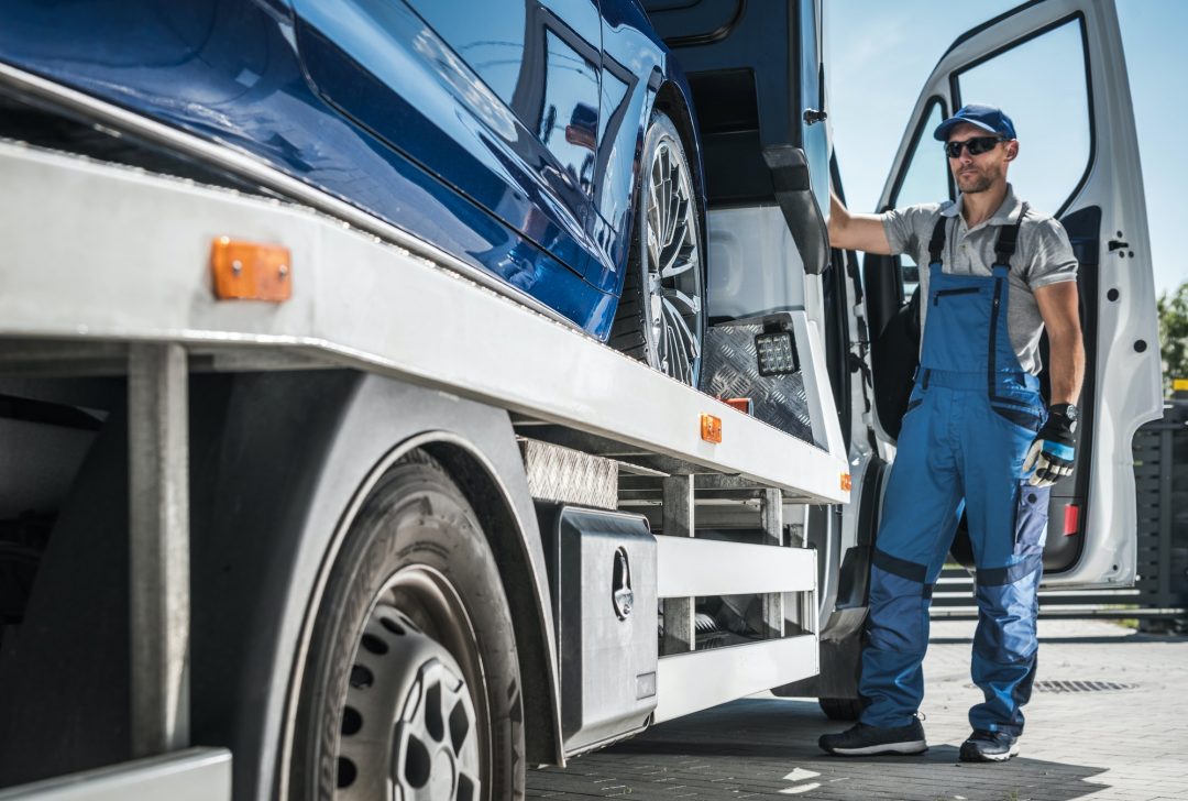 A tow truck driver in blue overalls and cap secures a blue car onto a flatbed truck. The sunny day highlights the reflective car surface and professional demeanor.