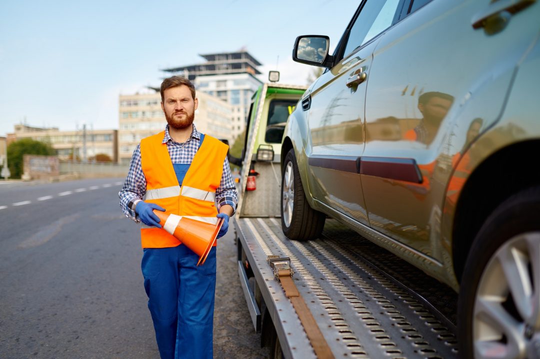 A tow truck driver in a reflective vest and gloves stands beside a flatbed tow truck, holding an orange traffic cone. A car is secured on the truck.